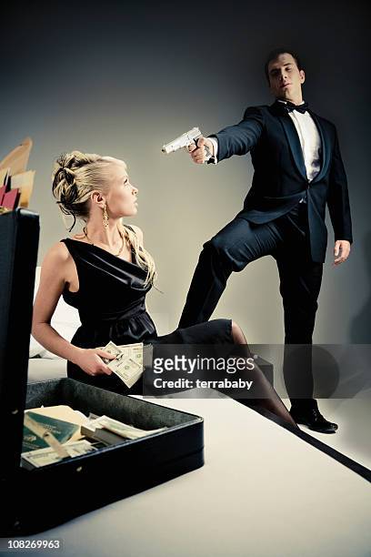 young spy man holding gun to woman looking through suitcase - female gangster stock pictures, royalty-free photos & images
