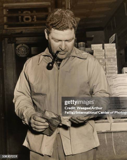 Man, smoking a pipe, holds two packages in front of a diary store, March 1943. Stacks of Velveeta, Kraft, and Borden processed cheeses are visible...