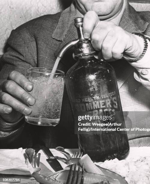 Man fills a glass from a bottle of 'Good Health Seltzer, Sommer's Beverages, Registered Conents 26 Oz New York,' in the foreground is a plate of...