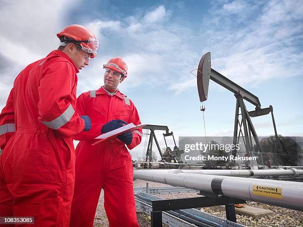 two workers looking at a clipboard in the foreground with onshore oil pumps above oil wells (nodding donkeys/pumpjacks) in the background - working oil pumps stock pictures, royalty-free photos & images