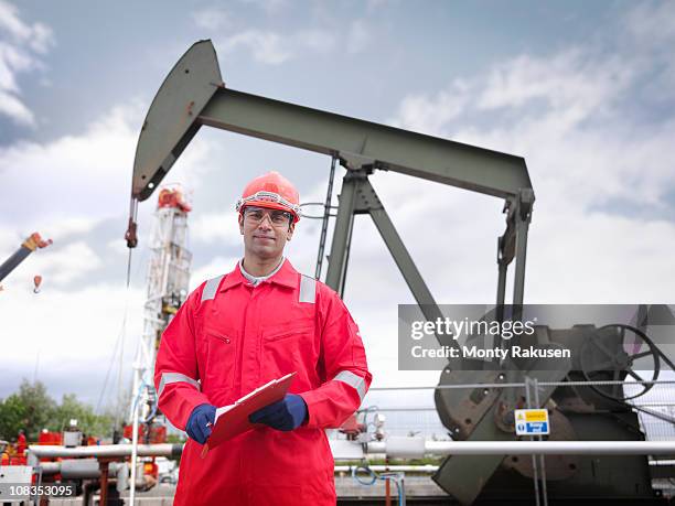worker looking to camera, standing in front of an oil pump above an onshore oil well (nodding donkey/pumpjack) - red jumpsuit stock pictures, royalty-free photos & images