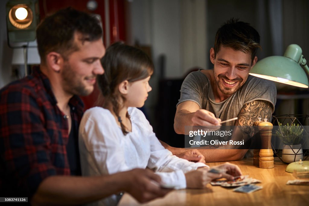 Two men playing cards with girl at table at home