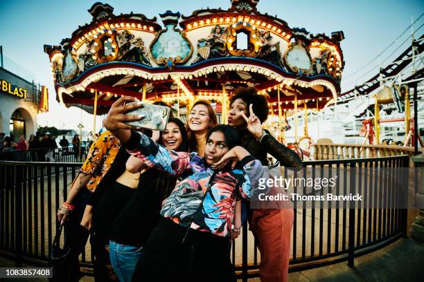 Female friends taking selfie with smart phone in front of carousel at amusement park