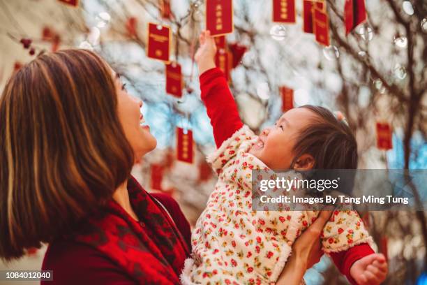 pretty young mom holding her lovely toddler girl to reach the red envelopes in chinese new year joyfully. - chinese new year stock pictures, royalty-free photos & images