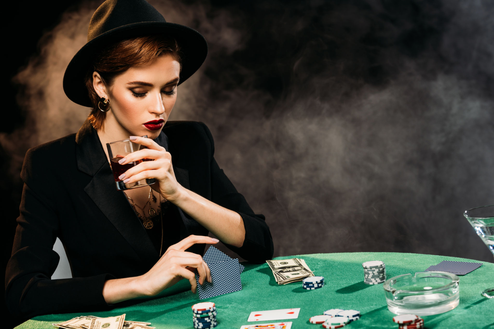 pensive attractive girl in jacket and hat playing poker and holding glass of whiskey in casino pensive attractive girl in jacket and hat playing poker and holding glass of whiskey in casino