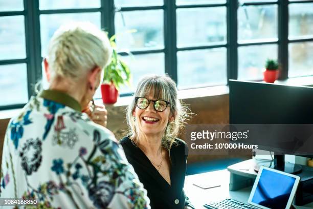 cheerful mature woman at desk smiling towards senior female colleague - friends chatting mature foto e immagini stock