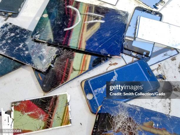 Close-up of a large number of broken cellphones, including cracked cellphone screens with exposed wiring, on a white surface, suggesting E Waste...