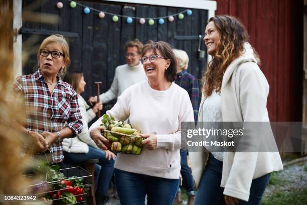 cheerful women standing with baskets against friends at farm during dinner party - community events fotografías e imágenes de stock