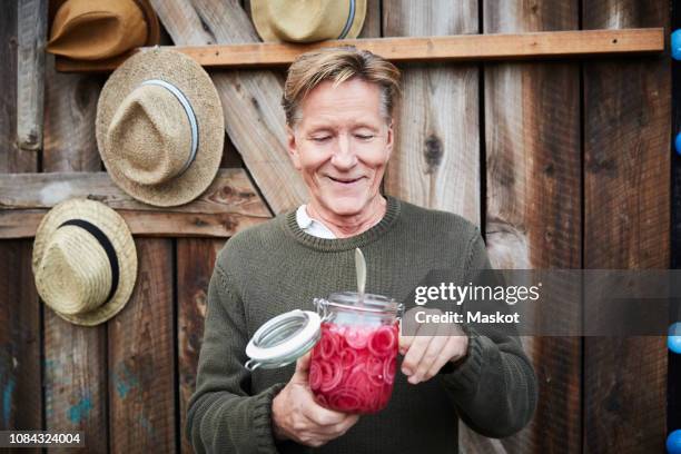 smiling senior man holding pickled onion jar against barn door - pickle jar stockfoto's en -beelden