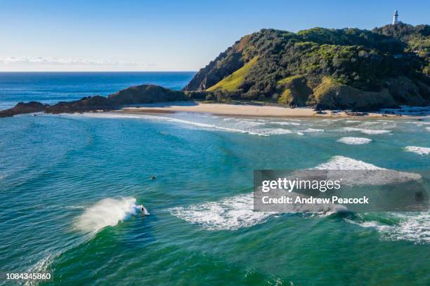 a surfer catches a wave at wategos beach below cape byron lighthouse which sits on australia's most easterly point near byron bay - byron bay hinterland stockfoto's en -beelden