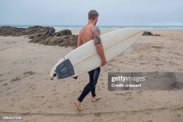 hombre caminando con tabla de surf en la playa de fistral, newquay, cornwall en un día de otoño. - newquay fotografías e imágenes de stock