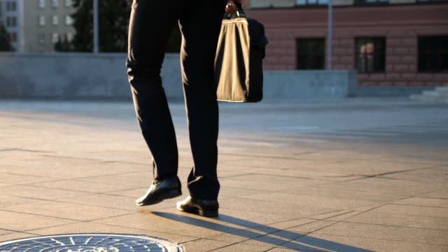 Feet of young businessman with a briefcase walking in city street. Business man commuting to work. Confident guy in suit being on his way to work. Cityscape background. Slow motion Rear view Close up