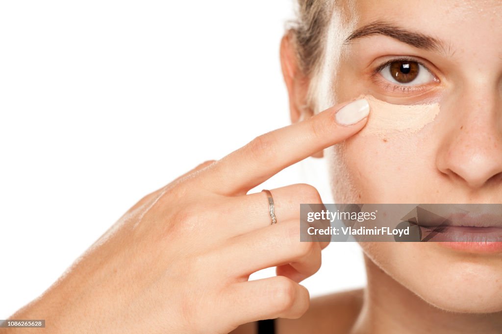 Young woman applying concealer under her eyes on white background