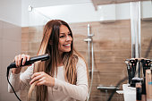 Beautiful young smiling woman using a hair straightener while looking into the mirror in bathroom.
