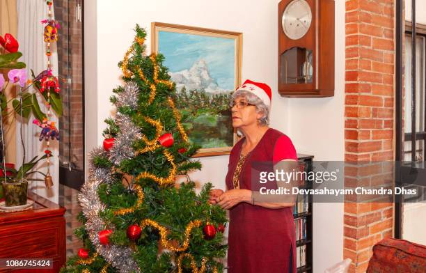 a grey haired senior latin american lady with a christmas cap is seen decorating an artificial christmas tree with great concentration, for the family in her living room.; she is now adding christmas ornaments - woman fingers in ears stock pictures, royalty-free photos & images