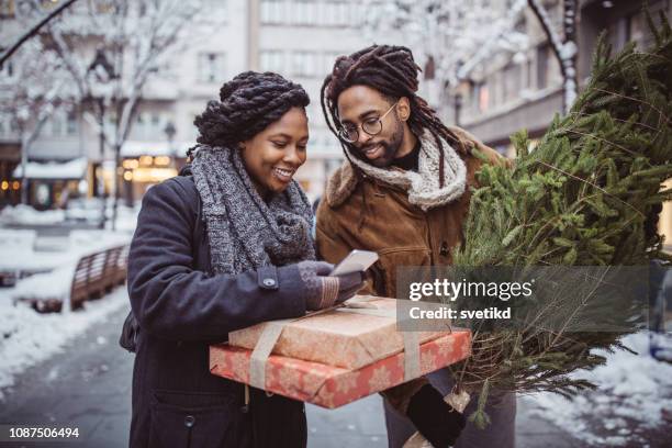 jong koppel lopen de straten van de stad, holding geschenkdozen en kerstboom - african people buying a christmas tree stockfoto's en -beelden