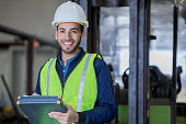 Young Hispanic man working as forklift operator in large warehouse smiles while wearing safety gear