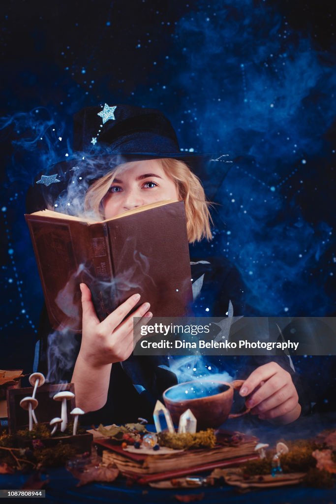 Charming young witch with a book of spells and magical potions. Magical workplace with candles, mysterious smoke, crystals and mushrooms. Dark background with stars and copy space