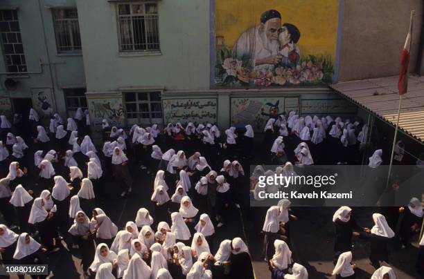 High-angle view of primary school girls in traditional headscarves as they talk in groups in the school's courtyard under a mural of Ayatollah...