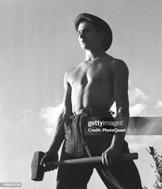 Young member of the Civilian Conservation Corps stands shirtless while holding a sledge hammer, ca 1930s. The CCC was a work relief program which was...