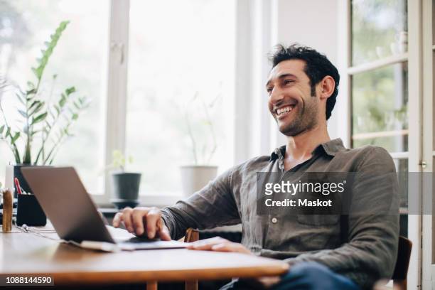 happy businessman using laptop at table by window in home office - looking away stock-fotos und bilder