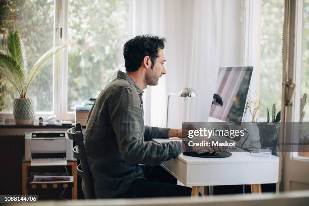 side view of young businessman using computer while sitting at desk in home office - pc home stock-fotos und bilder