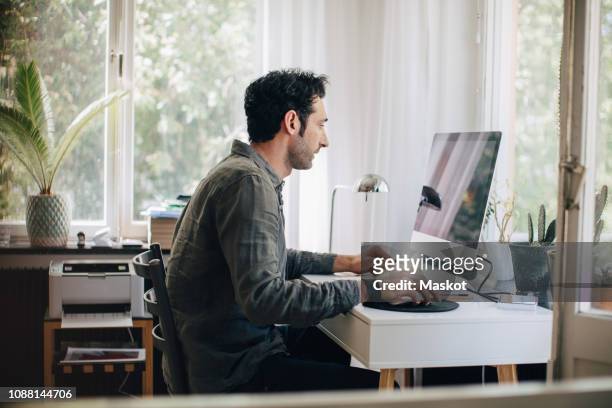 side view of young businessman using computer while sitting at desk in home office - teleworking icon stock pictures, royalty-free photos & images