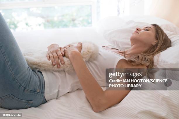 young woman on bed with hot water bottle on tummy - woman holding tummy stock-fotos und bilder