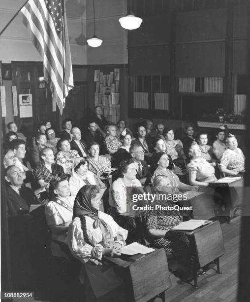Underneath an American flag, a group of immigrants sit in a classroom while learning English for their citizenship, early to mid twentieth century.