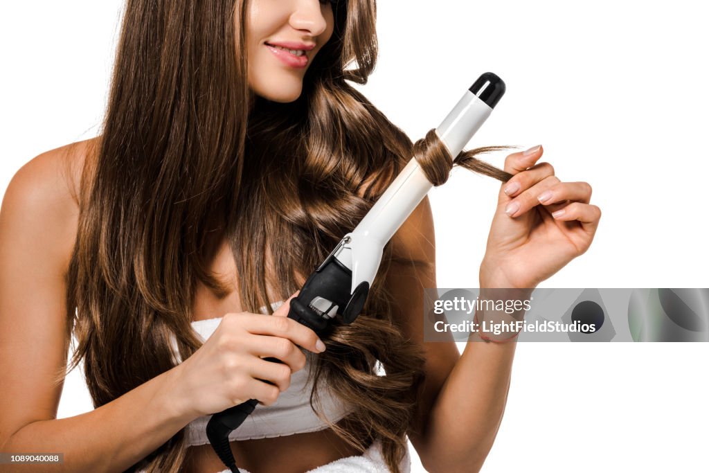 Cropped view of girl with long brown hair using curling iron isolated on white