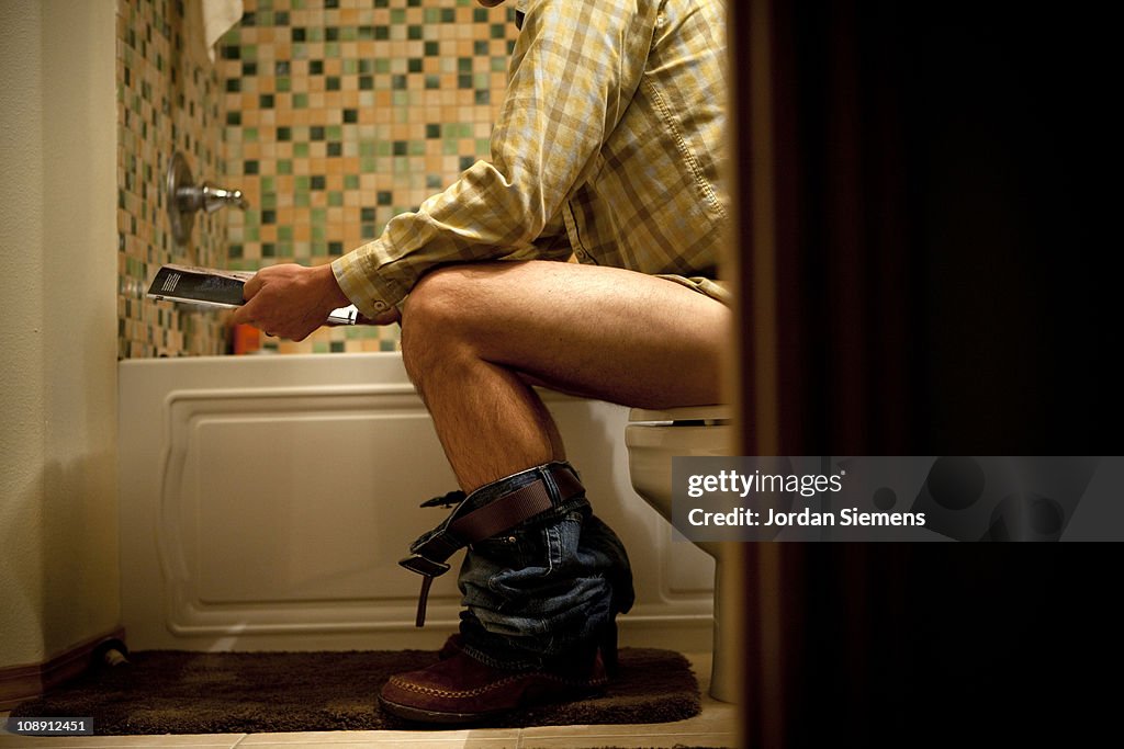 Male reading magazine on toilet.