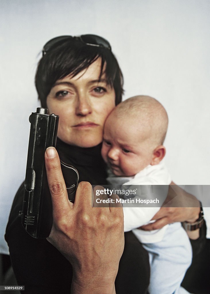 A mother holding a handgun up and a baby in her other arm