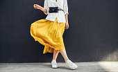Horizontal cropped image of stylish slim woman in beautiful yellow skirt. Caucasian female fashion model standing over gray wall background outdoor with copy space.