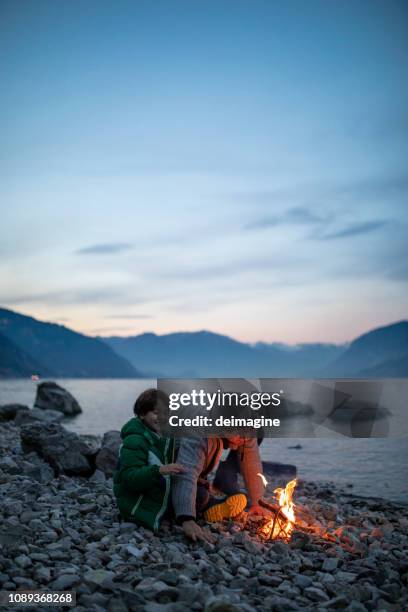 father and son light a fire together in front of the lake - bonfire beach stock pictures, royalty-free photos & images