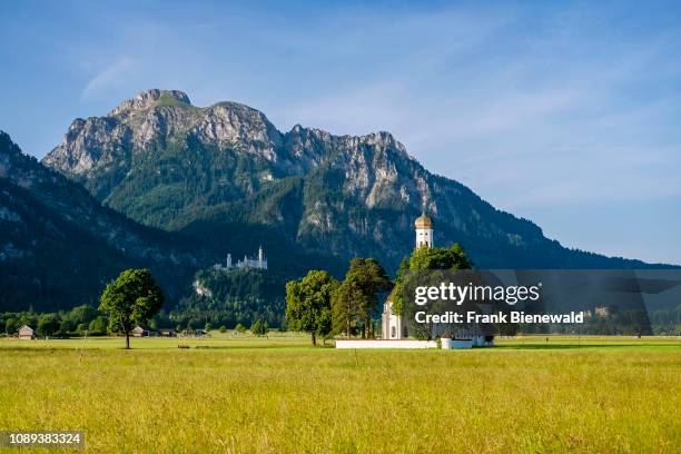 The church St. Coloman, surrounded by trees, the alps and castle Neuschwanstein in the distance.