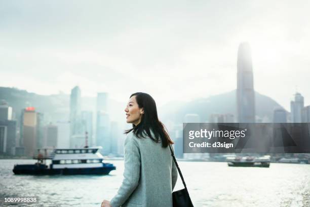 confident young lady looking forward to start her day on a fresh bright morning while standing against hong kong cityscape - prosperity stock pictures, royalty-free photos & images