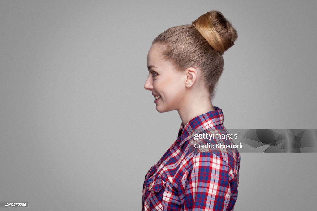 Profile side view of beautiful blonde girl in red, pink checkered shirt, collected bun hairstyle standing and looking aside with toothy smile.