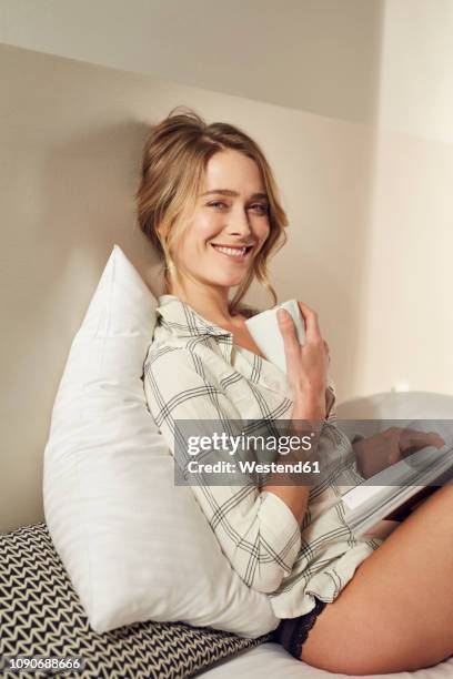 portrait of smiling woman sitting on bed with coffee mug and magazine - women in slips stock-fotos und bilder