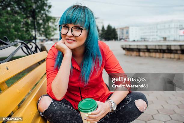 portrait of smiling young woman with dyed blue hair sitting on a bench with beverage - spinner stock-fotos und bilder