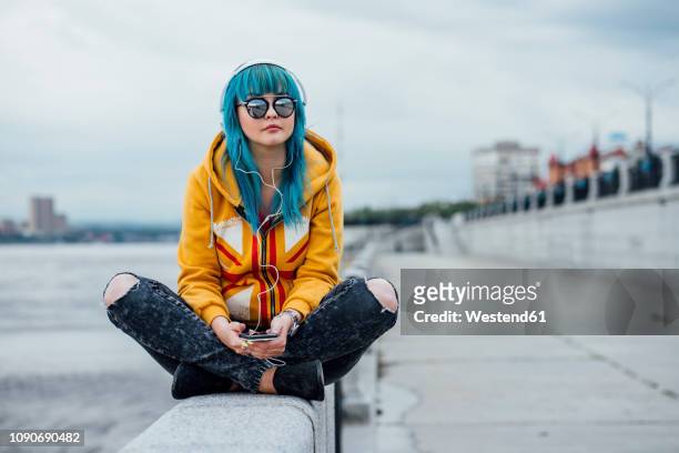 young woman with dyed blue hair sitting on a wall listening music with headphones and smartphone - dyed shades stock pictures, royalty-free photos & images