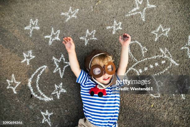 portrait of smiling toddler wearing pilot hat and goggles lying on asphalt painted with airplane, moon and stars - kinderwunsch stock-fotos und bilder