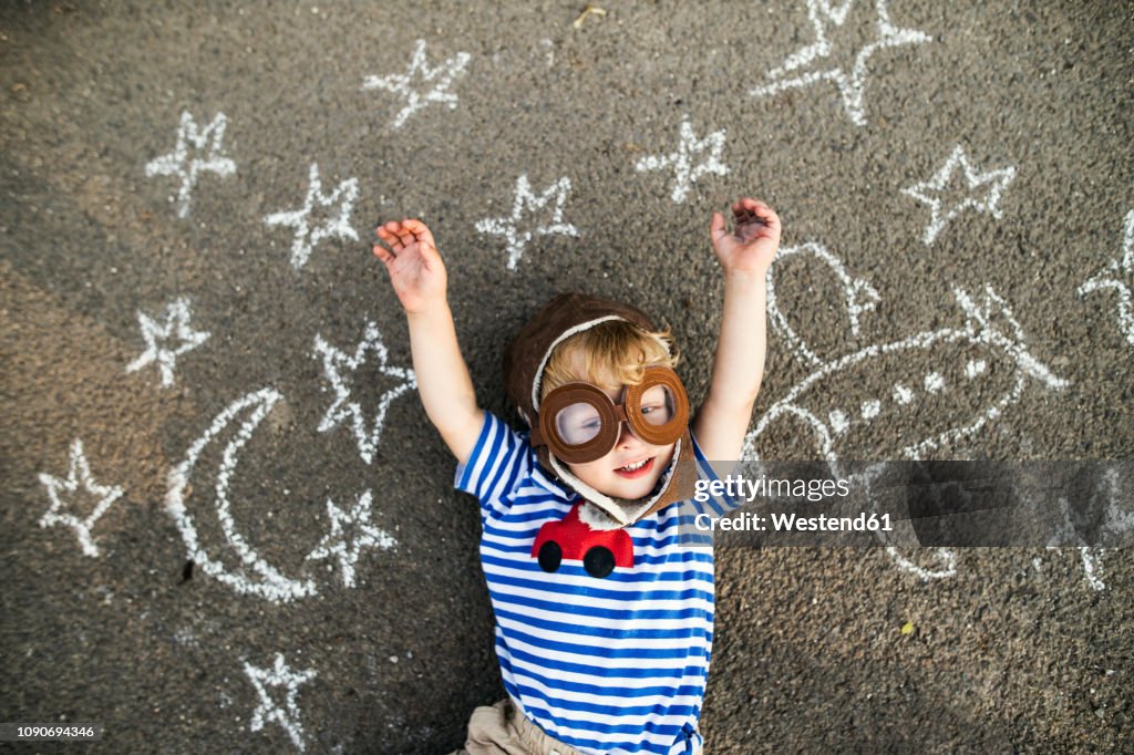 Portrait of smiling toddler wearing pilot hat and goggles lying on asphalt painted with airplane, moon and stars