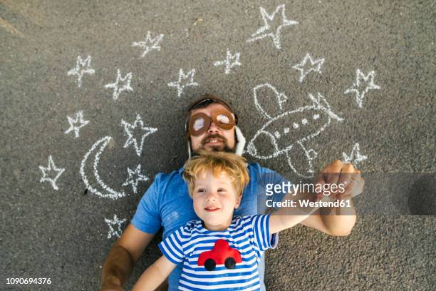 portrait of mature man wearing pilot hat and his little son lying on asphalt painted with airplane, moon and stars - family drawing 個照片及圖片檔
