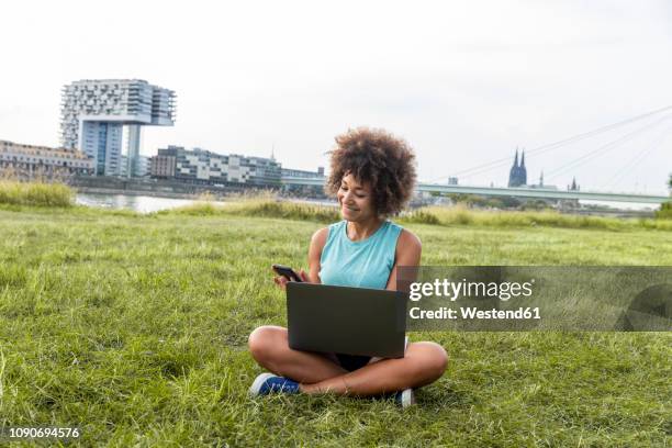 germany, cologne, woman sitting on meadow using laptop and cell phone - woman using smartphone with laptop stock-fotos und bilder