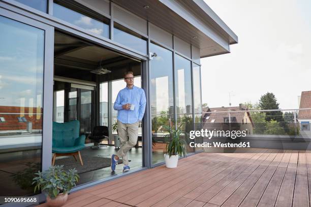 young man with cup of coffee standing at balcony door - dachterasse stock-fotos und bilder