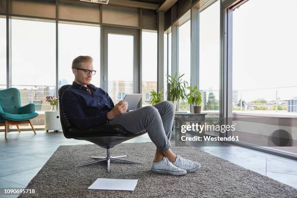 young man sitting in armchair at the window at home using tablet - capital architectural feature - fotografias e filmes do acervo