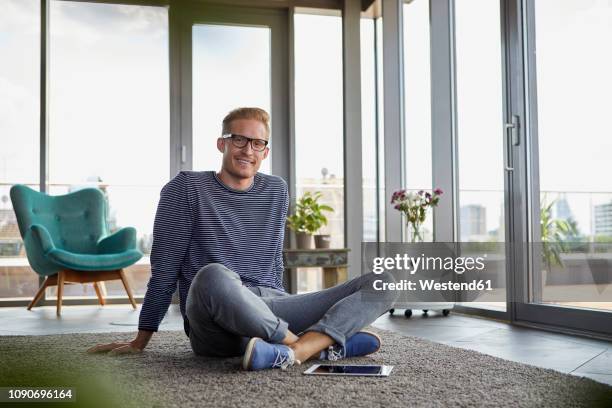 portrait of smiling young man sitting on carpet with tablet at home - sitting on floor fotografías e imágenes de stock