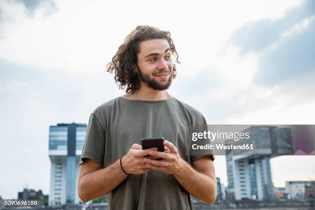 germany, cologne, smiling young man holding cell phone looking sideways - at a glance foto e immagini stock