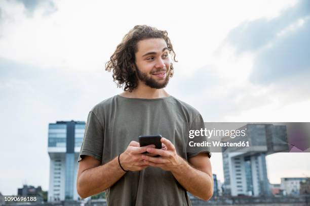 germany, cologne, smiling young man holding cell phone looking sideways - sideways glance - fotografias e filmes do acervo