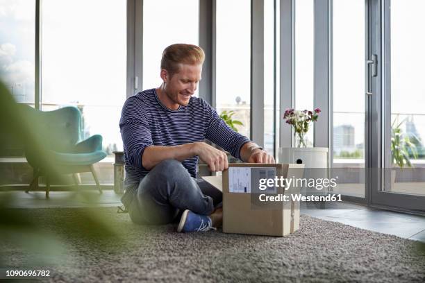 smiling young man sitting on carpet at home unpacking parcel - man shopping happy stock pictures, royalty-free photos & images